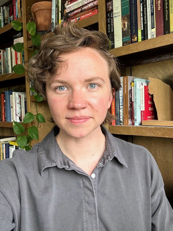 A photo of a white person in front of a shelf of books and plants. They have short curly hair and a nose ring and are wearing a black polo.